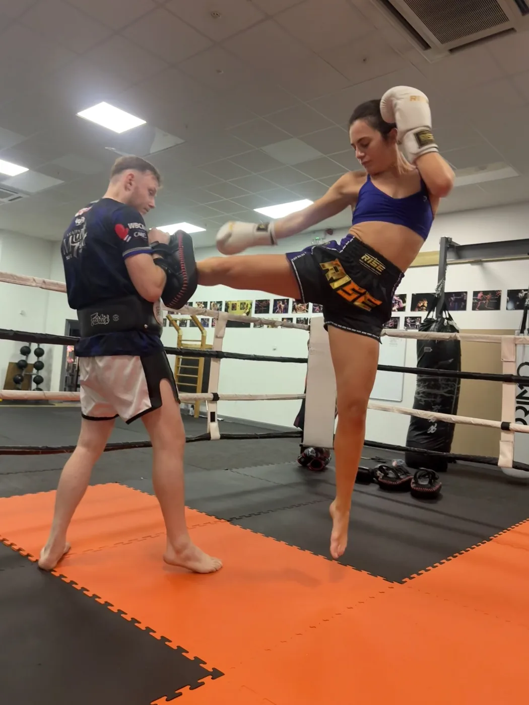 A fighter throwing a high kick into a trainer's pads inside a gym ring